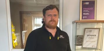 Man in a black polo shirt with a logo on the chest standing in a doorway.