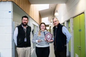 Three people standing in a hallway, a woman holding a plaque.