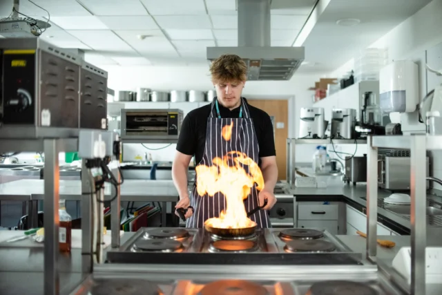 a chef standing in a kitchen with flames coming up from a pan