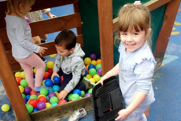 Three children playing in ball pit, one girl smiling with a bag