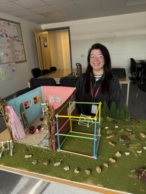 A young woman standing behind a model of a house and play area with a climbing frame.