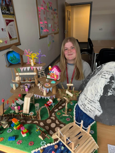 A young girl smiling behind a table with a miniature wooden village and forest diorama.
