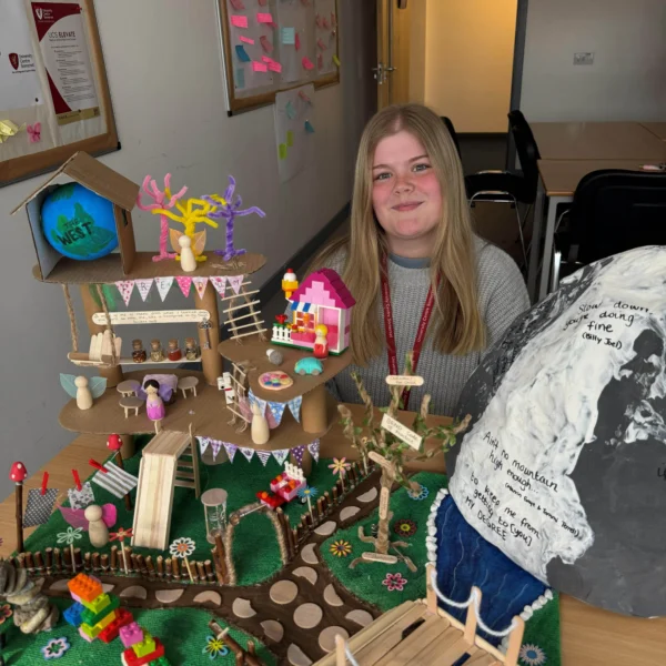A young girl smiling behind a table with a miniature wooden village and forest diorama.