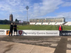 Somerset County Cricket Club field with a church tower in the background and UCS students proudly standing beside the UCS perimeter advertising board at the ground