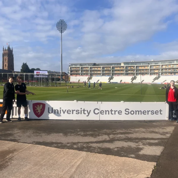 Somerset County Cricket Club field with a church tower in the background and UCS students proudly standing beside the UCS perimeter advertising board at the ground
