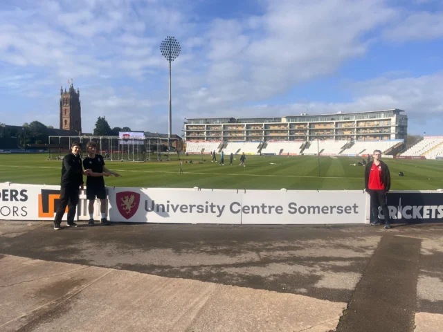 Somerset County Cricket Club field with a church tower in the background and UCS students proudly standing beside the UCS perimeter advertising board at the ground
