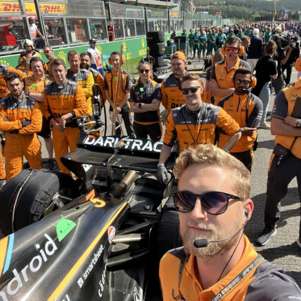 Joel Vermiglio takes a selfie with the Mclaren Formula 1 race team and race car in the pit at a grand prix.