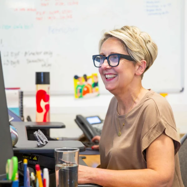 A woman with short blonde hair and glasses sitting at her desk in front of a computer monitor.
