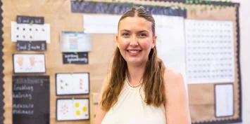 A young woman with long brown hair wearing a white sleeveless top standing in front of a classroom bulletin board.