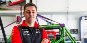 A smiling man in a racing team jacket standing in a garage.