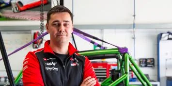 A smiling man in a racing team jacket standing in a garage.