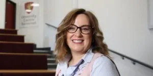 A smiling woman with brown hair and glasses standing in front of a staircase.