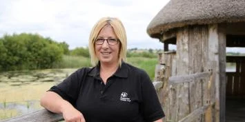Woman with blond hair wearing a black polo shirt standing next to a wooden structure.