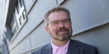 A man with short hair and glasses standing in front of a building with a blurred background.