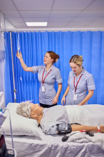 Two nurses standing over a patient in a hospital bed, one holding an IV pole. The patient is lying down with a blood pressure cuff on their arm.