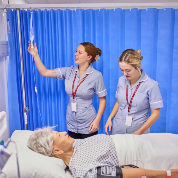 Two nurses standing over a patient in a hospital bed, one holding an IV pole. The patient is lying down with a blood pressure cuff on their arm.