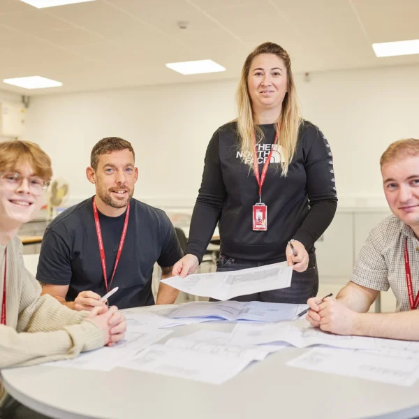 A woman stands behind a table with three seated students, all wearing red lanyards, looking at papers.