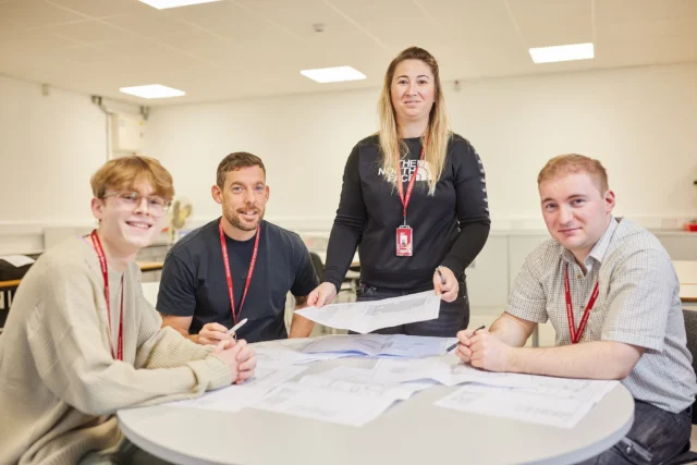 A woman stands behind a table with three seated students, all wearing red lanyards, looking at papers.