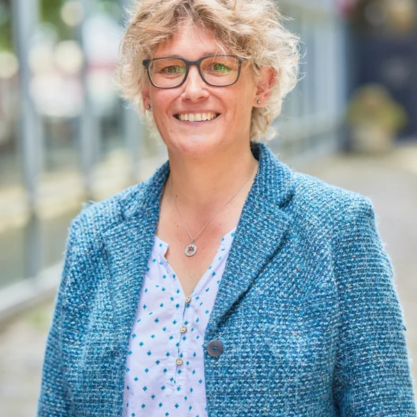 A woman with short blonde hair and glasses smiling outside, wearing a blue blazer and a white blouse with blue dots.