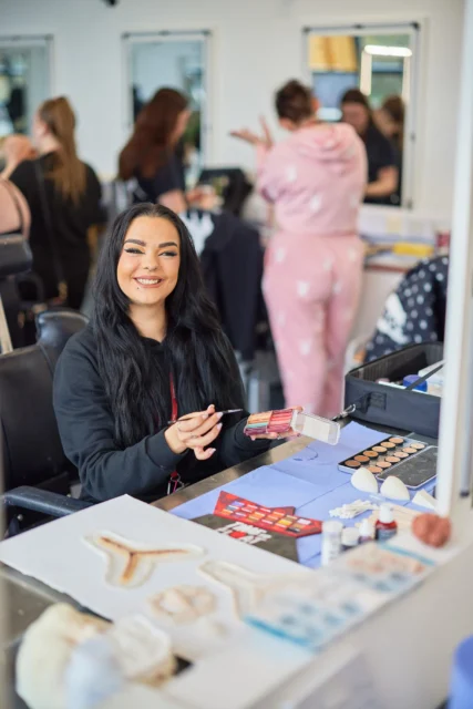 A woman with long black hair sitting at a table with makeup products in a busy salon.