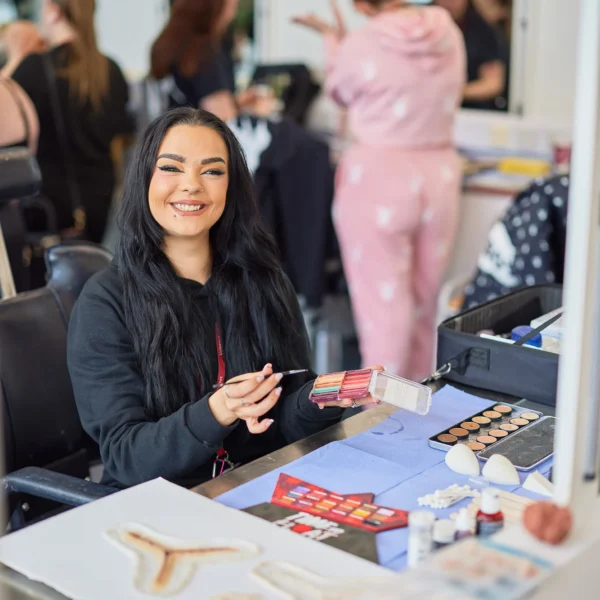 A woman with long black hair sitting at a table with makeup products in a busy salon.