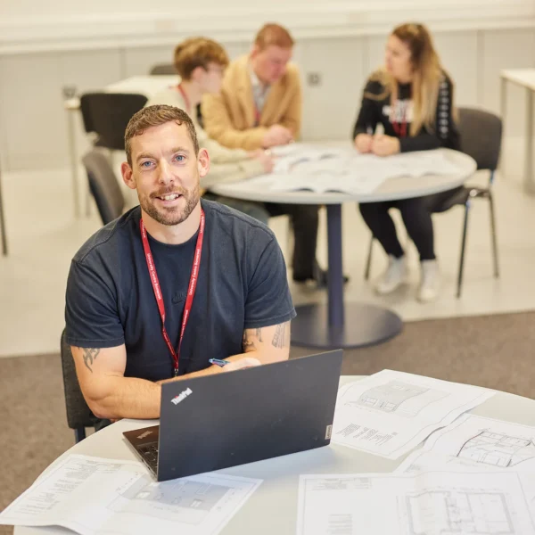 Man sitting at a desk with a laptop and some papers, wearing a red lanyard around his neck.