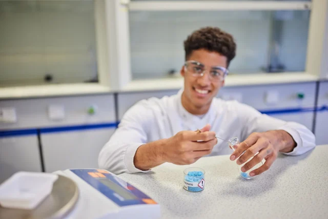 Young man in a lab coat and safety goggles handling a small object.