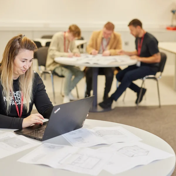 A woman studies on her laptop with scattered papers on the table. She sits at a table in a classroom with other students in the background.