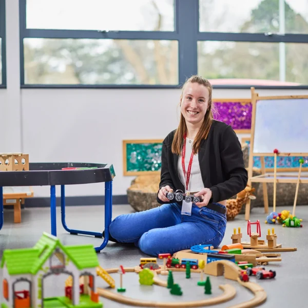 Woman sitting on floor with toy train set, smiling at camera in classroom setting.