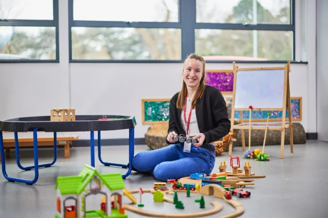 Woman sitting on floor with toy train set, smiling at camera in classroom setting.