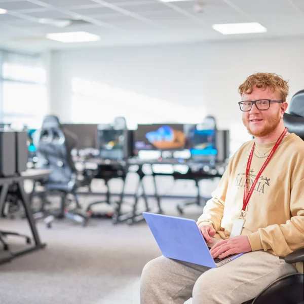 A smiling man holding a clipboard sits on a gaming chair in a room with computers.