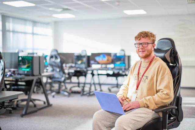 A smiling man holding a clipboard sits on a gaming chair in a room with computers.