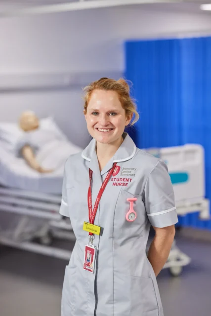 A smiling nurse standing in a hospital room with a mannequin on a bed.