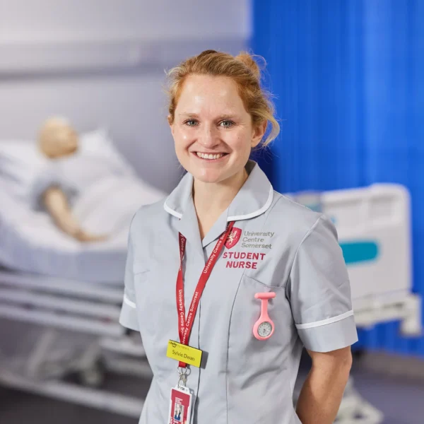 A smiling nurse standing in a hospital room with a mannequin on a bed.