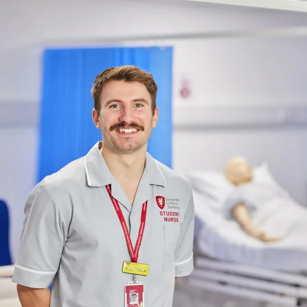 A smiling man in a light grey hospital top with a red lanyard in a hospital room with a blue curtain.