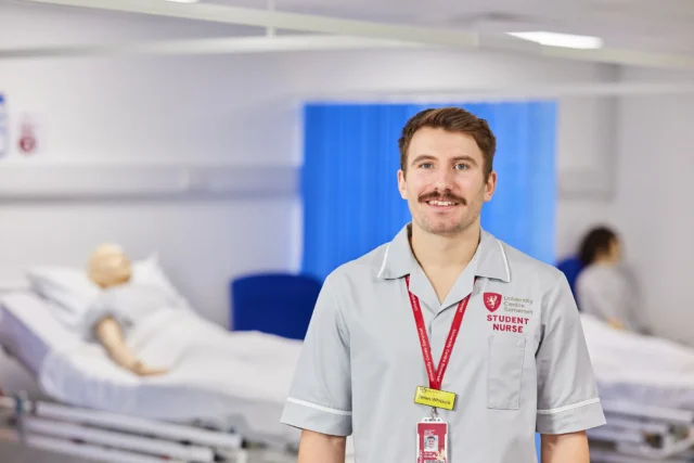 A man in a medical uniform standing in front of a hospital bed with a mannequin.