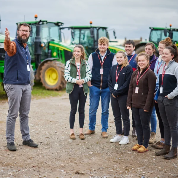A man in casual clothing gestures while standing in front of a group of people, with large green tractors behind them.