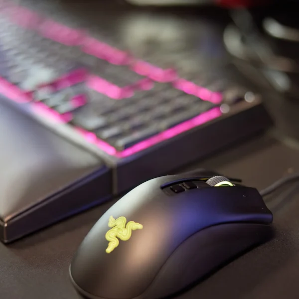 Black computer mouse with a yellow logo next to a keyboard with pink backlighting.