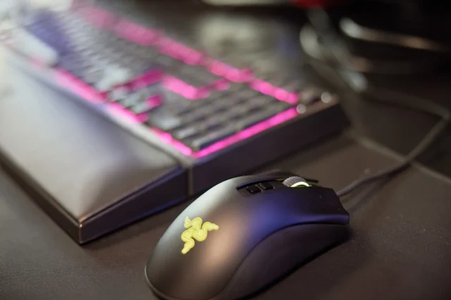 Black computer mouse with a yellow logo next to a keyboard with pink backlighting.