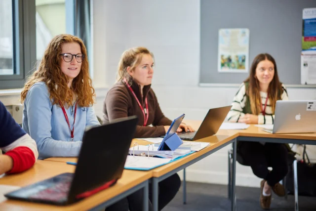 Three women sitting at desks with laptops in a classroom setting