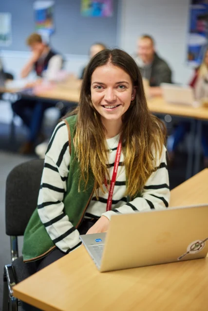 Young woman sitting at a desk with a laptop in a classroom setting.