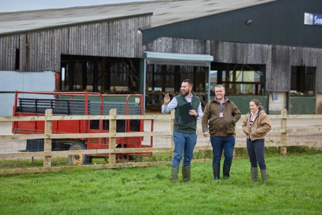 Three people standing in a grassy field in front of a barn, smiling and chatting.
