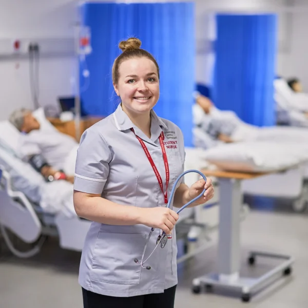 A smiling nurse standing in a hospital ward with patients in beds.