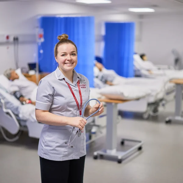 A smiling nurse standing in a hospital ward with patients in beds.