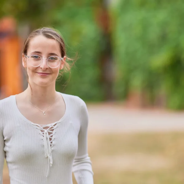 A young woman smiling outdoors in a park with trees and grass.