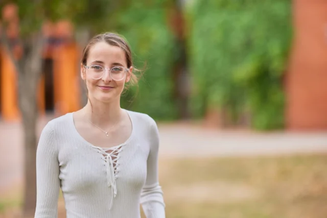A young woman smiling outdoors in a park with trees and grass.