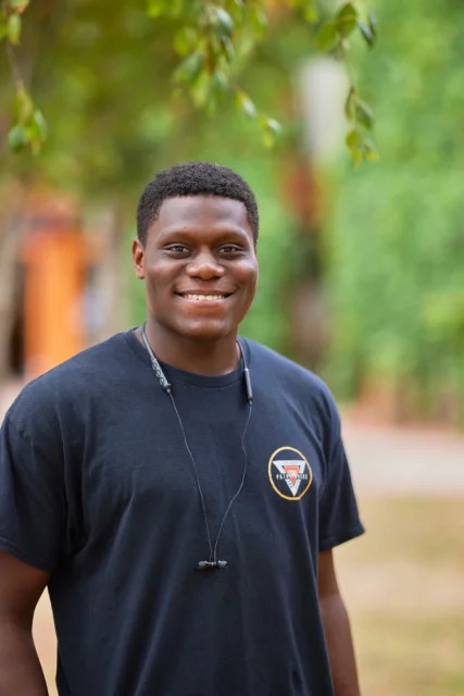 A young man with short black hair wearing a black T-shirt and a whistle around his neck.