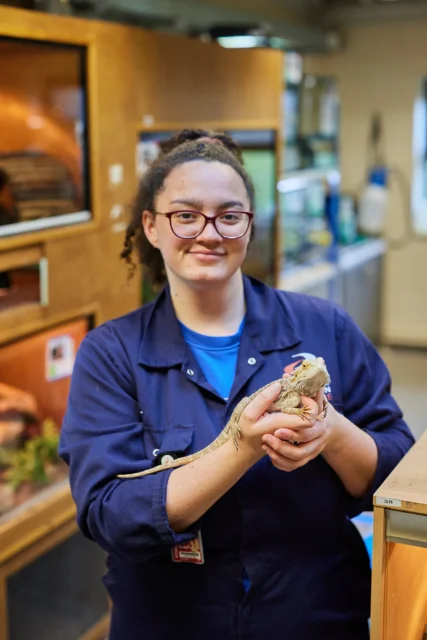 A woman wearing a blue jumpsuit holding a lizard in her hands.