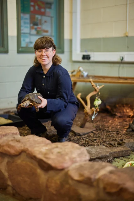 A woman crouches down holding a lizard in her hands.
