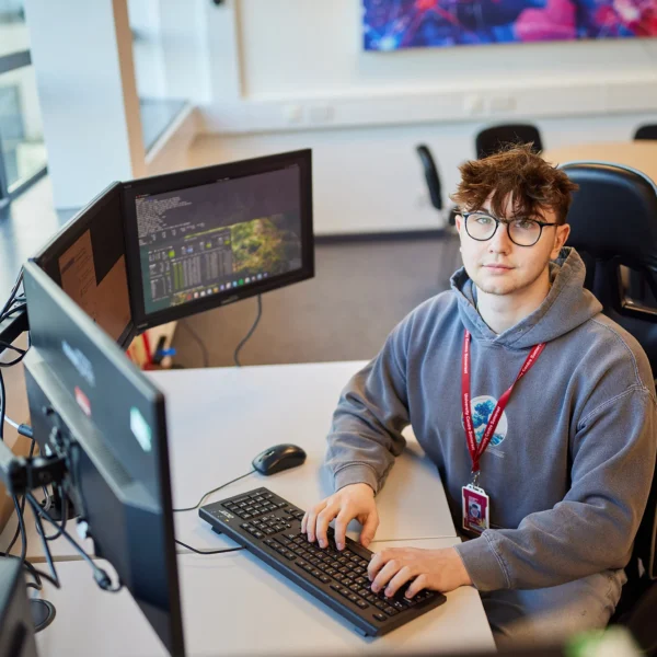 A young man sits at a desk with two computer monitors, focused on work.